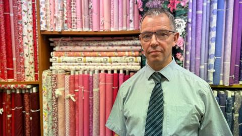 A man with glasses, shirt & tie standing in front of a selection of colourful fabrics.