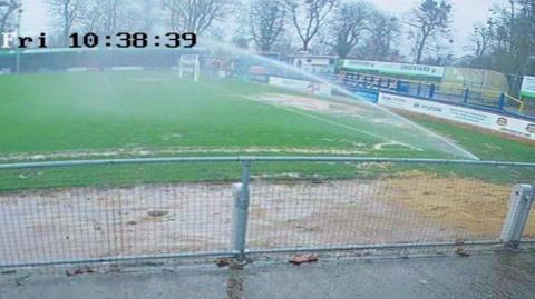 A CCTV image of half of an empty football pitch with water spraying from a sprinkler in the corner quadrant. Spectator areas are on the far side of the pitch and on the right.