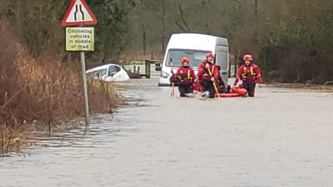 Three rescue workers can be seen wading through water at knee height - a submerged van and car can be seen in the background