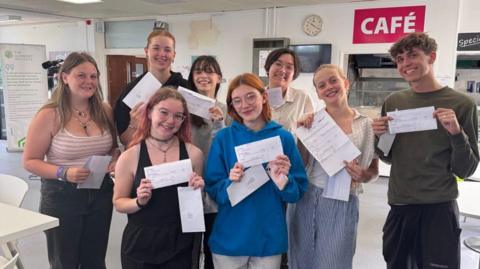 Eight students, holding up their results. They are in a room with a white wall and grey floor. They are all smiling. 