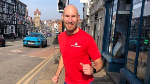 A man with a red t-shirt, beard and a shaved head looks at the camera and is smiling. His hands are in a running position and behind him are shops and a road with cars on it. 