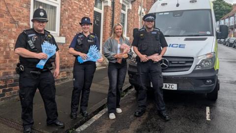 Three uniformed police officers - a man and a woman - and another woman wearing jeans and a grey fleece sanding in front of a marked police van parked on a terraced street. The two on the left are holding blue hand-shaped leaflets with printing on them and the other two leaflets depicting hairy hands.