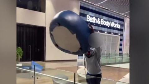 A screenshot from a video, showing an obscured person holding a heavy blue padded seat over the edge of a glass barrier in Westfield Shopping Centre