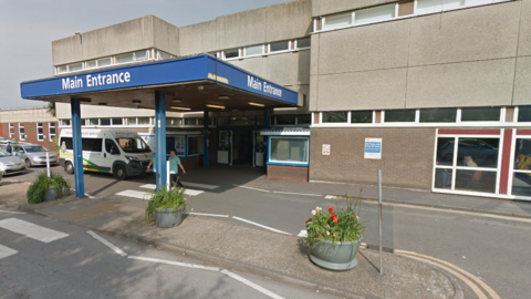 An exterior picture of Eastbourne District General Hospital - the main building is grey and the entrance is below a blue roof which has Main Entrance displayed at the front. 