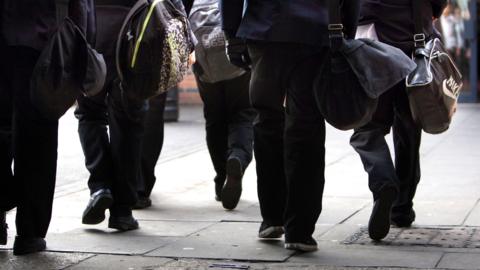 A ground-level stock image of children / young adults walking away from the camera carrying satchels