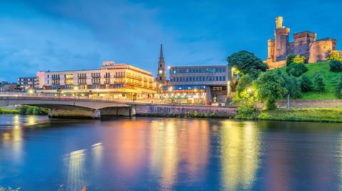 Buildings on Inverness' riverside are lit up at night. The buildings include a large block of flats, the city centre's steeple and Inverness Castle. The lights are reflected in the River Ness.