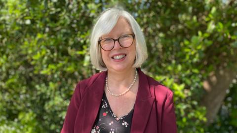 Dr Nicola Brink stands outdoors on a sunny day, wearing a burgundy jacket and glasses, with greenery in the background behind them.