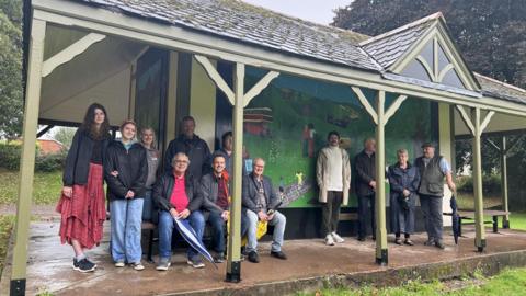 Local residents are pictured sitting on benches and standing in front of the park shelter. The building is a rectangular shape and has a pointed roof over the top of it. The green pattern can be seen on the shelter wall.