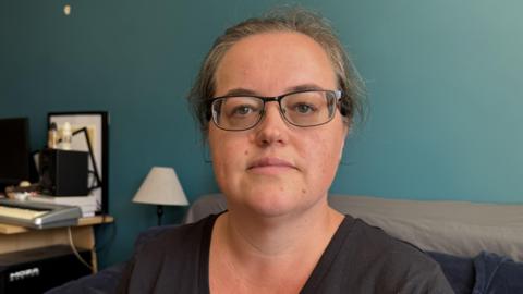 A woman sits on a dark blue sofa in a room with teal-colored walls. She has her hair pulled back and she is wearing glasses and a black V-neck shirt. Behind her, there is a desk setup with computer equipment, including a monitor and speakers, along with shelves holding various items. A small lamp is placed on the desk, and another piece of furniture is partially visible to the left of the person.