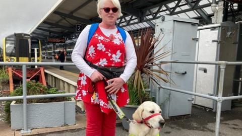 blind woman wearing red jeans standing with golden labrador guide dog at train station