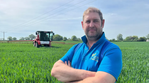 A man with short blonde hair and a beard standing in a field in a blue polo shirt with long green shoots all around him and a red tractor in the background