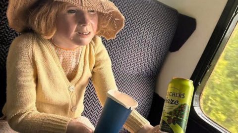 A child mannequin with a straw hat and cardigan sitting on a train. The mannequin is holding a can. The train appears to be moving.