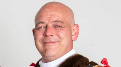A headshot photo of a man wearing mayoral clothes. He has a bald head and is smiling at the camera.