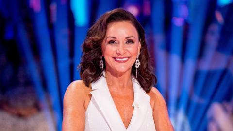 Shirley Ballas wearing a white dress sitting in the judge's chair on the BBC One's Strictly Come Dancing set 