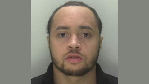 A police custody photo of Kiano Houghton Jones. He has short dark brown hair, a dark brown beard and is wearing a black collared top.