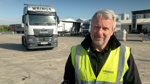 A man stood in a yard with a large white lorry behind him. He is wearing a reflective yellow jacket. He has short grey hair and a short beard.