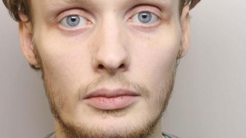 A man with a thin beard and moustache and blue eyes stares at the camera. He is against a grey background