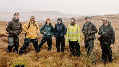 Five men and two woman wearing waterproof outerwear, smiling and standing on open grassland.