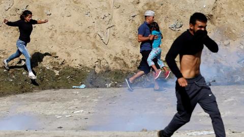 Migrants and children covering mouths with tear gas cloud behind