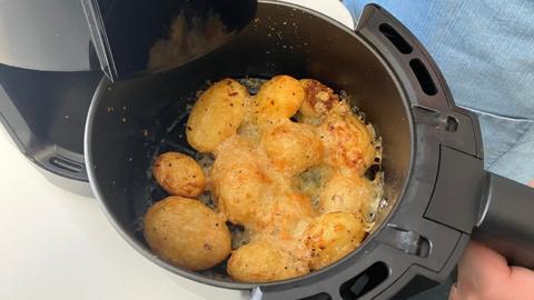An air fryer basket filled with golden-brown roasted potatoes, topped with melted cheese or seasoning. A person is holding the basket, which is partially open to reveal the contents.