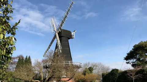 An old fashioned windmill stands against a blue sky, surrounded by trees.