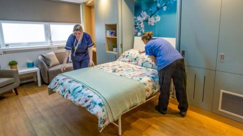 Two midwives prepare a bed with a floral bedspread in a comfortable looking room