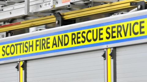 A close-up of the side of a fire engine. The name "Scottish Fire and Rescue Service" is in black letters on a yellow background.