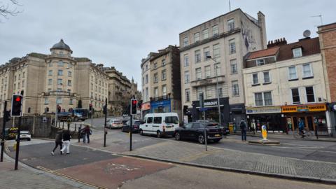 The junction where Anchor Road, College Green and St Augustine's Parade meet in Bristol.  Pedestrians are crossing Anchor Road, while cars wait at lights on the other side of the road. The Marriott Hotel can be seen in the distance - and there are several nearby takeaways. 