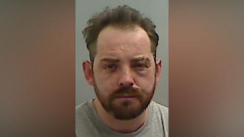 Mugshot of Sean Miller. He has a short ginger beard and short messy dark hair. He has a cut on his nose and swelling and bruising around his left eye.