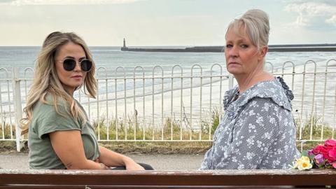 Two women sit on a bench overlooking the sea