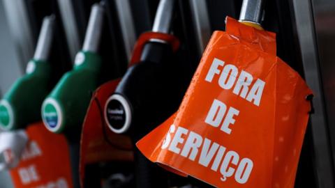 A close-up photograph shows fuel pump nozzles docked in their pumping station. One is wrapped in an orange sign reading "For de Servico" - the Portuguese for out of service