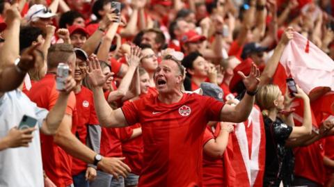 An image showing a Canada fan expressively celebrating a Canada goal against Qatar in the stands with a friend. He is wearing a red Canada jersey. Around him are dozens of people wearing red and white, waving Canadian flags and smiling. 