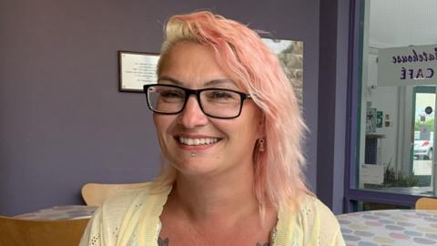 Charlotte is sitting in a cafe, wearing a yellow top. She has pink hair and wears glasses.