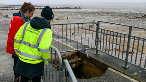 Two women looking at a fenced-off sinkhole on the edge of a promenade. The women are on the left. Beyond the sea wall is a beach and the sea.