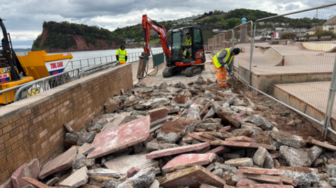 Construction workers in high-visibility clothing break up a concrete walkway beside a brick wall at a coastal site, using a small excavator and tools, with piles of rubble in the foreground and the sea and hillside buildings in the background.
