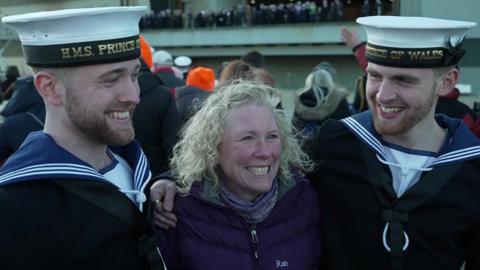 Vicky Mackinnon stands in the middle of her sons, Alex and Callum. They all have their arms around each other and are grinning. Callum (left) and Alex (right) are both wearing their Royal Navy uniforms. Vicky has short curly blonde hair and wears a purple quilted puffer jacket with a purple scarf.