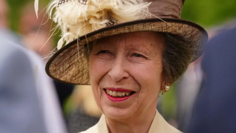 A close-up of Princess Anne smiling. She had short brown hair which is tucked under a brown hat.
