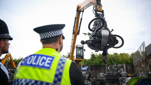 Two police officers watch as a scrap yard telehandler carries an assortment of e-bikes and scooters to a crusher