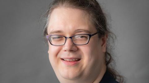Headshot of Chris Hilliard  - they are wearing a black jacket and black rimmed glasses with long hair in a ponytail