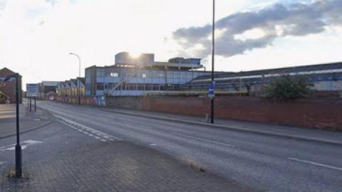 A wide, empty street in an urban area under a partly cloudy sky. The road curves gently and has clear lane markings, with pavements and signposts along the edges. Brick walls and industrial-style buildings line the right side of the street, while trees and smaller buildings appear on the left.