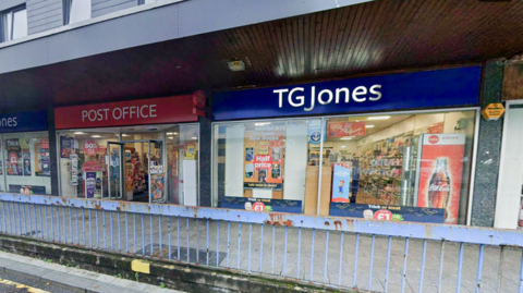A TG Jones shop with its name in white on blue background with a post office next door. There is a wide pavement in front and blue, rusted railings.