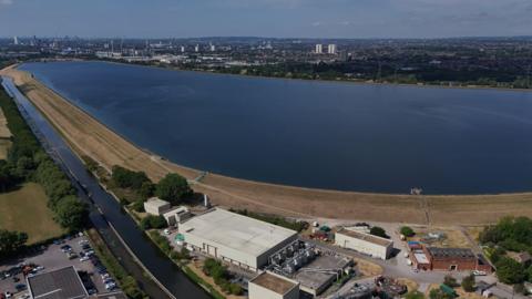 An image made with a drone shows the Chingford Reservoirs in London. A large body of water is surrounded by buildings in the foreground and trees in the background