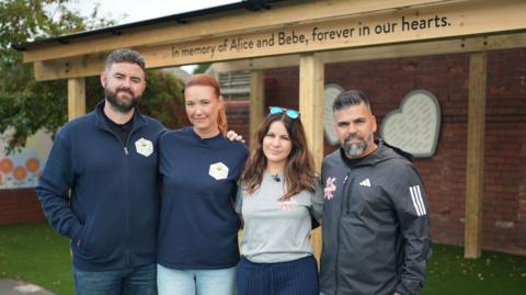Ben King, Lauren King, Alexandria Aguiar and Sergio Aguiar stand shoulder to shoulder in front of part of the new playground that has the words "In memory of Alice and Bebe, forever in our hearts."