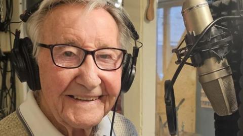 Mr Rayner looking at the camera and smiling. He has brown eyes and white hair. He is wearing headphones and is sitting in front of a microphone.