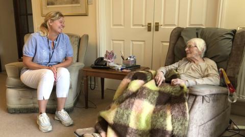 A white woman in a blue top and white trousers sitting in a chair next to an older woman with grey hair and a blanket over her knees