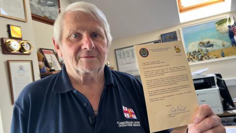 David Freeman looking at the camera holding a letter. He has short grey hair and is wearing a navy polo shirt with an RNLI logo on one side. Below the logo, it says "Flamborough lifeboat operations manager" in white capital letters. He is standing in an office. The walls are covered in art, accessories and awards.