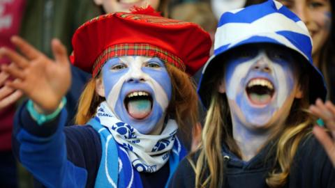 Two young girls in the crowd at a rugby match. Their faces are painted blue and white. One has a Saltire bucket hat, the other has a red tartan tammie hat. Their mouths are wide open and they are waving at the camera.