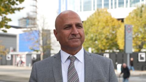 Councillor Naeem Ahktar stands in front of the former Ikea building in Coventry city centre. He is wearing a grey suit and has a grey tie with pink and white dots.