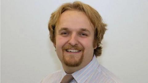 Robert Alden in a white shirt and brown tie. He has a goatee beard and dark blond hair.