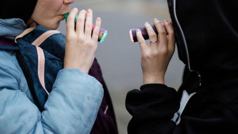 Children wearing winter jackets with hoods hold vapes in their mouths 